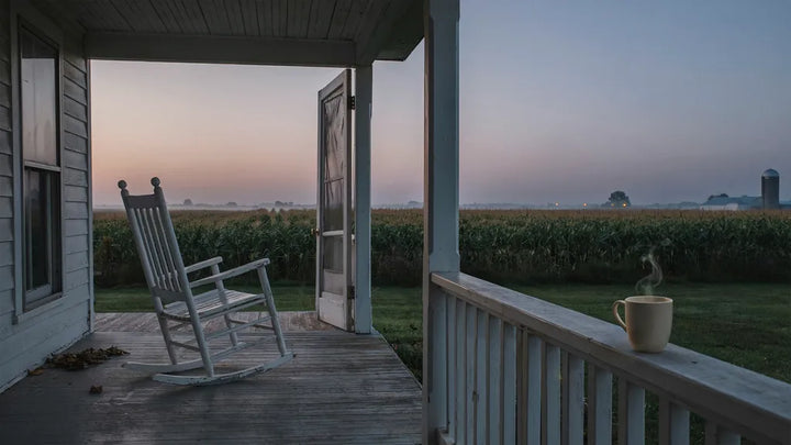 An empty rocking chair and a steaming coffee mug on a farmhouse porch railing at dusk — memorial song for grandpa's funeral