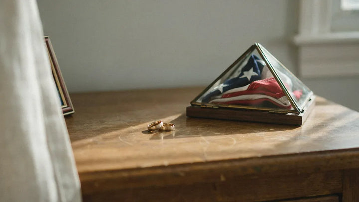 A folded flag in a display case beside two wedding rings on a nightstand in morning light