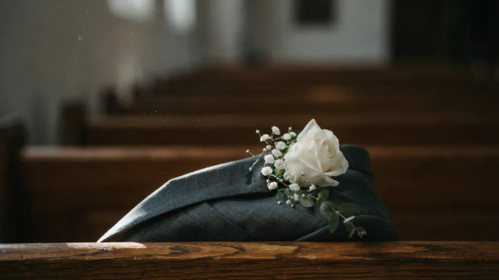 A white rose boutonniere pinned to a charcoal wedding suit lapel on a wooden church pew in soft morning light