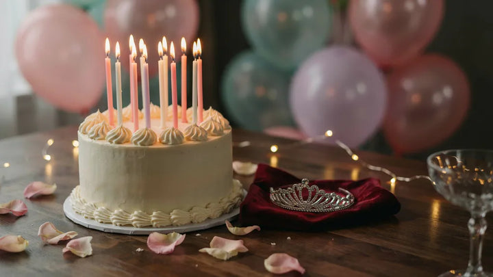 A candlelit birthday cake beside a small tiara on velvet, strung fairy lights glowing behind