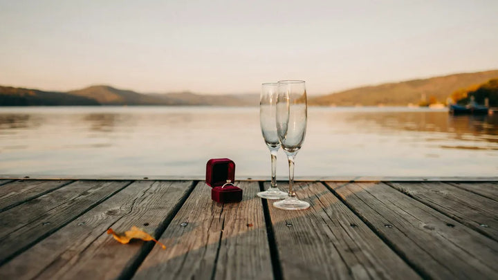 An open ring box with a diamond ring on a wooden lake dock at sunset, two champagne glasses beside it