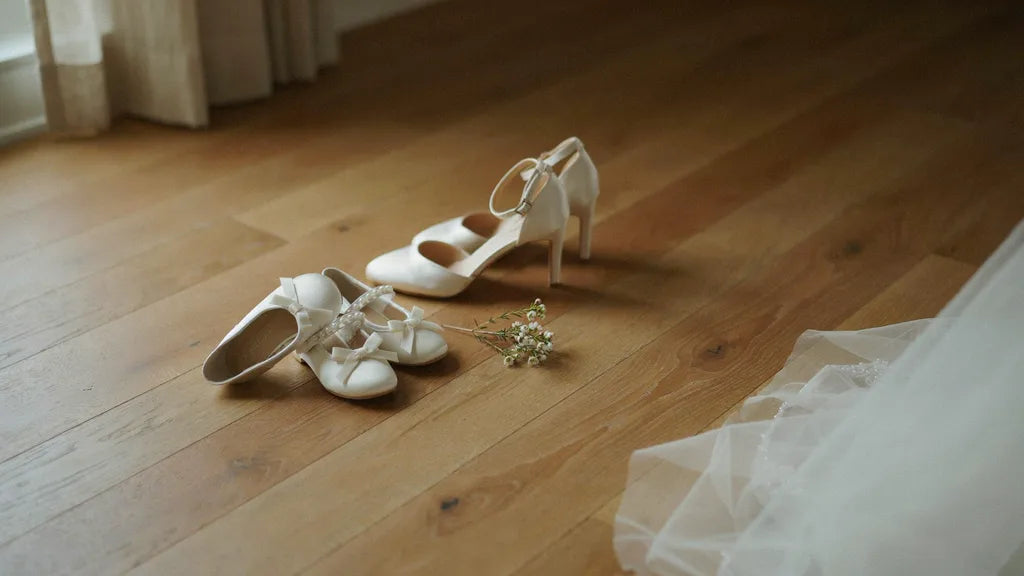 Tiny flower-girl shoes set next to adult wedding heels on a wood floor