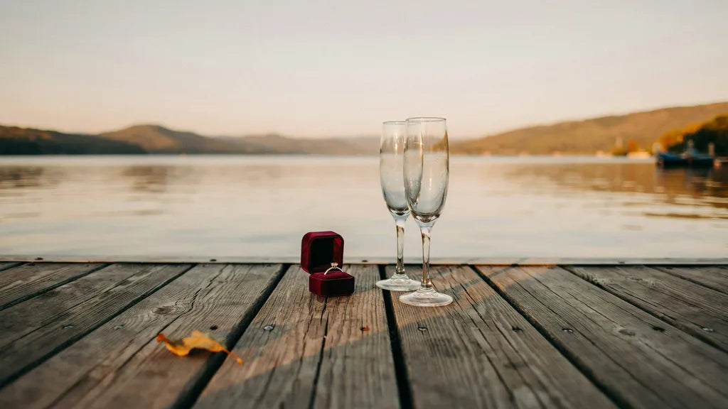 An open ring box with a diamond ring on a wooden lake dock at sunset, two champagne glasses beside it