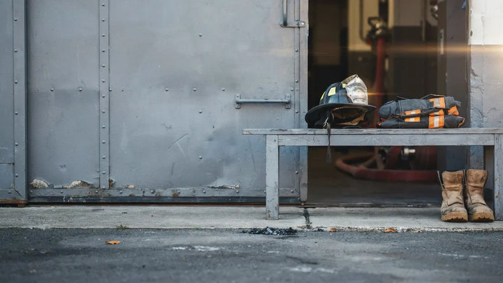 A firefighter's helmet, coat, and boots resting on a bench by a station door in soft morning light