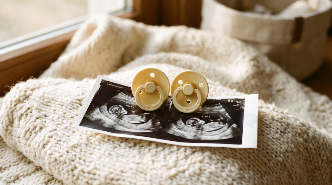 Two matching pacifiers resting side by side on a double sonogram printout on a cream knit blanket in warm window light