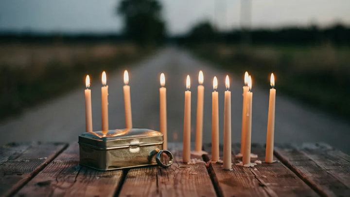 A tin keepsake box and ring beside ten lit candles on weathered wood with a country road behind