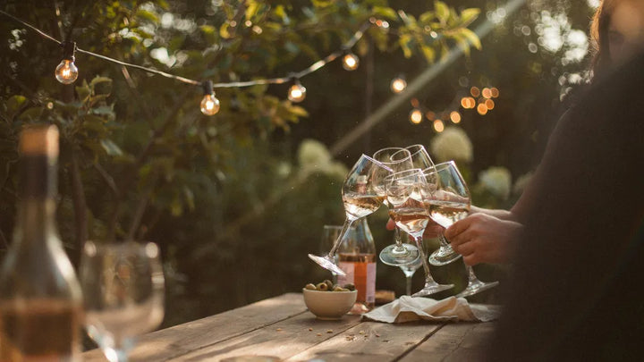 Raised glasses catching golden light over an outdoor table with warm string lights