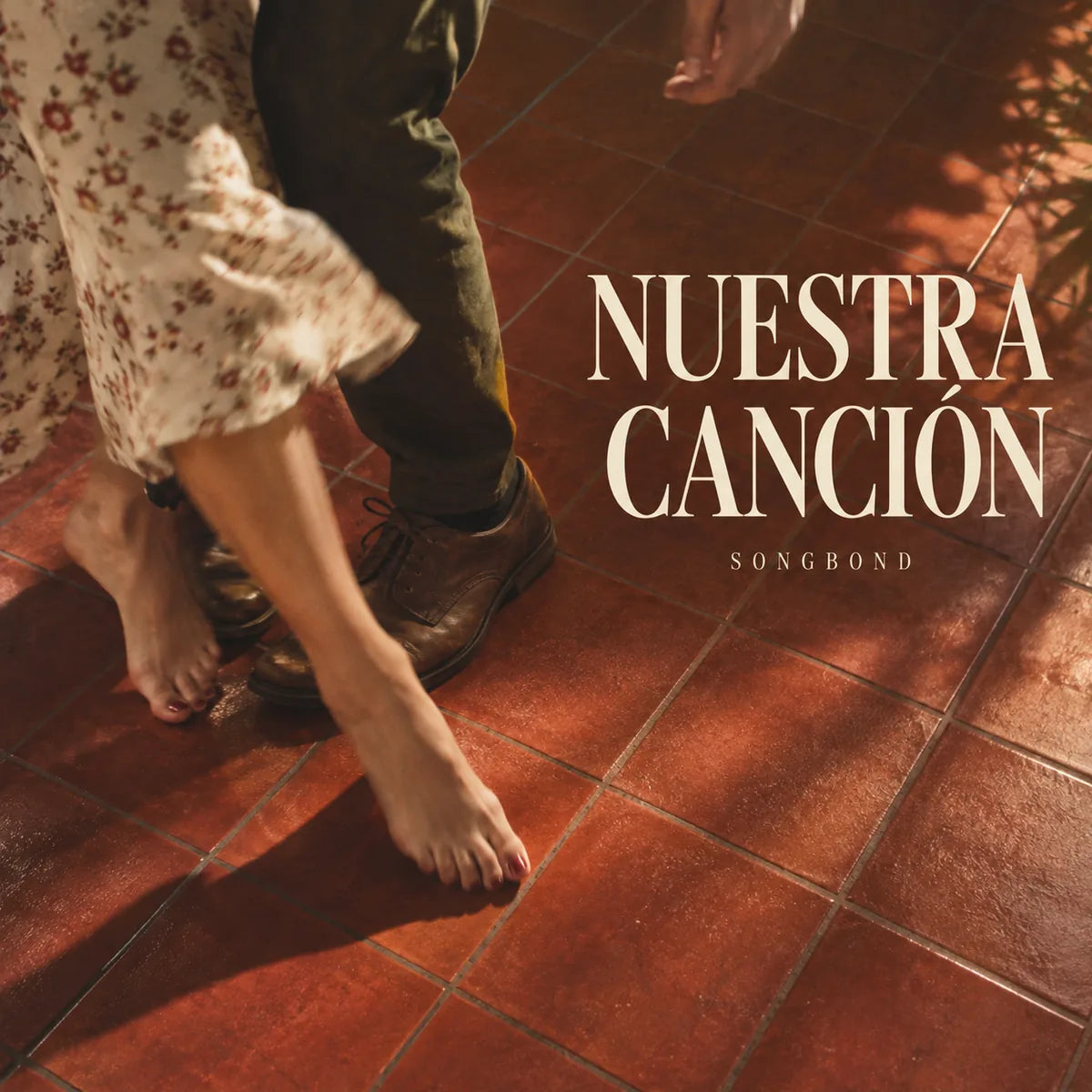 Music cover for Nuestra Canción by Songbond, with two adults' dancing feet on red kitchen tile beneath a large cream title with its Spanish accent.